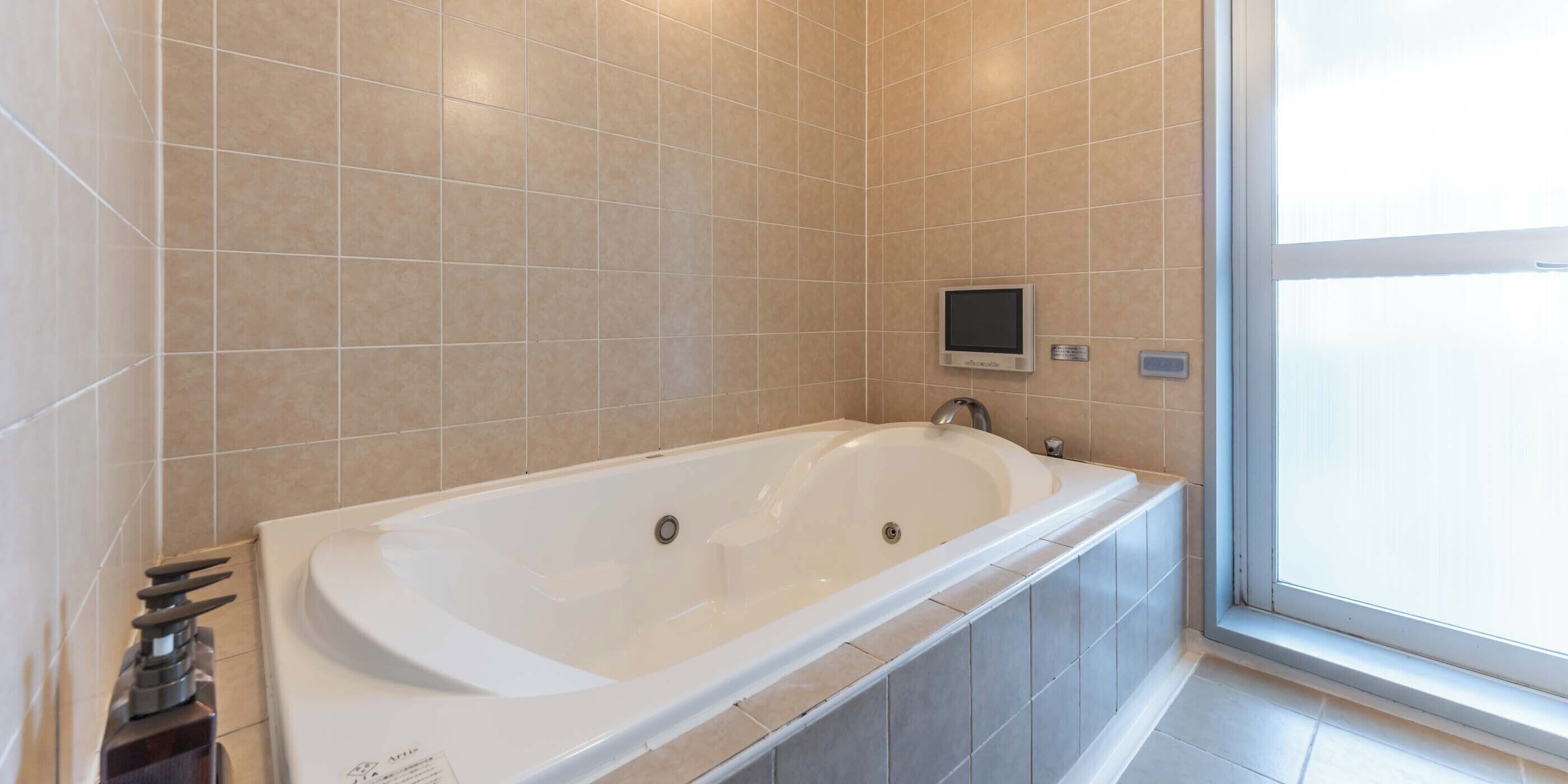 A modern bathroom with a large bathtub, tiled walls, a small screen, and a window.