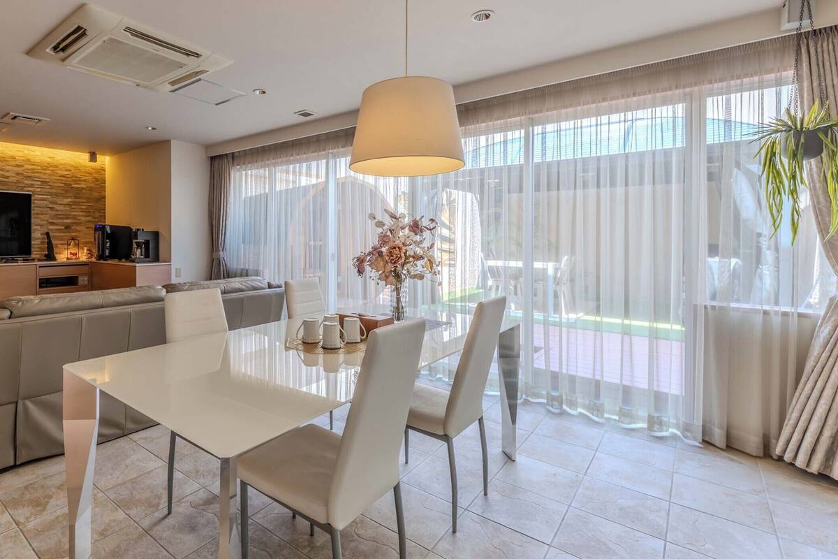 A modern living room with a TV, sofa, dining table, and large windows with sheer curtains.