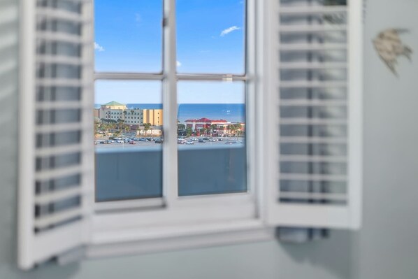 A window with white shutters, a view of a marina with boats, and buildings in the background. - South Harbour 8B - 𝑲𝒂𝒊 𝑴𝒂𝒌𝒂𝒏𝒊 (Pensacola Beach)