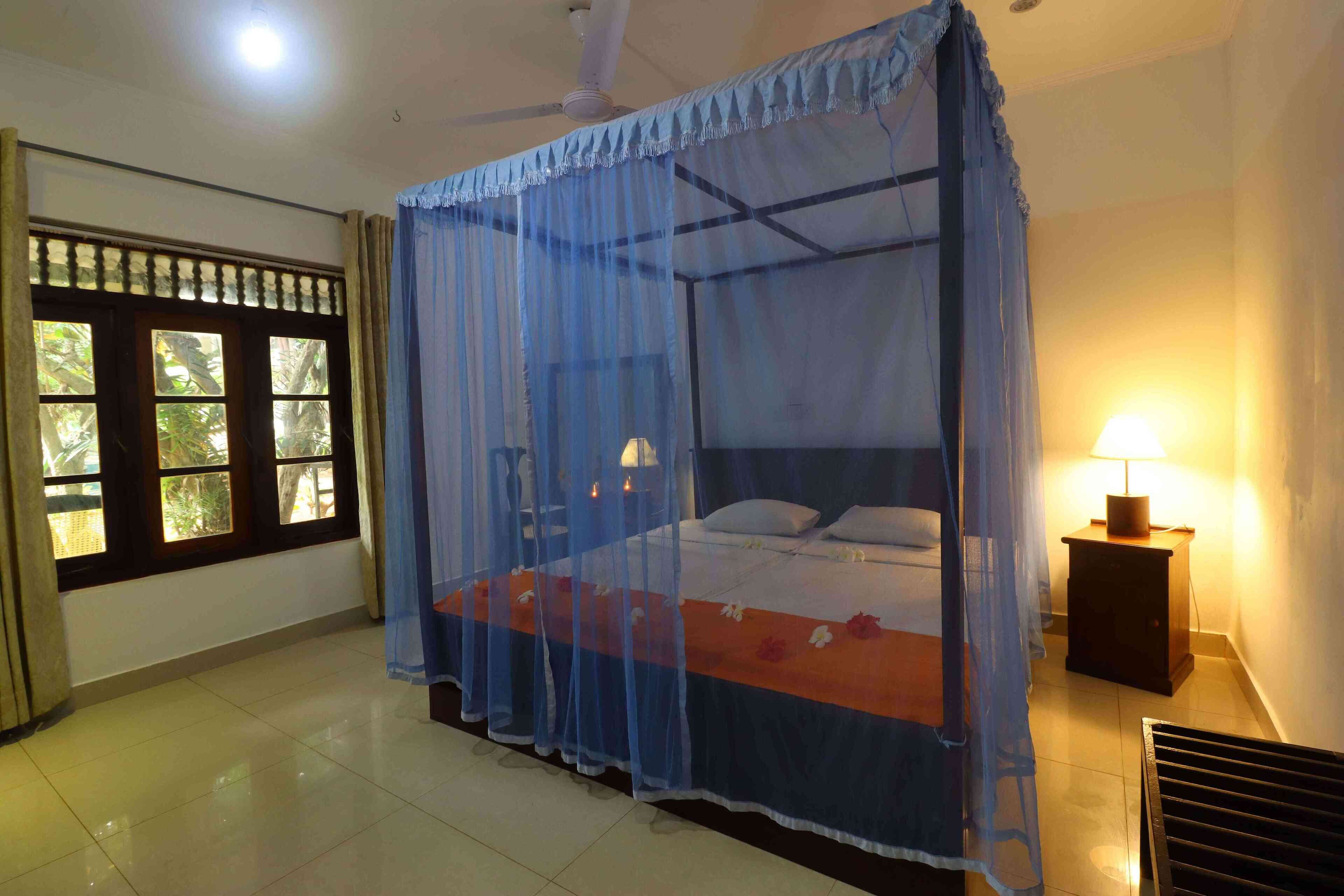 A room with a bed under a mosquito net, a nightstand with a lamp, and a window with curtains.