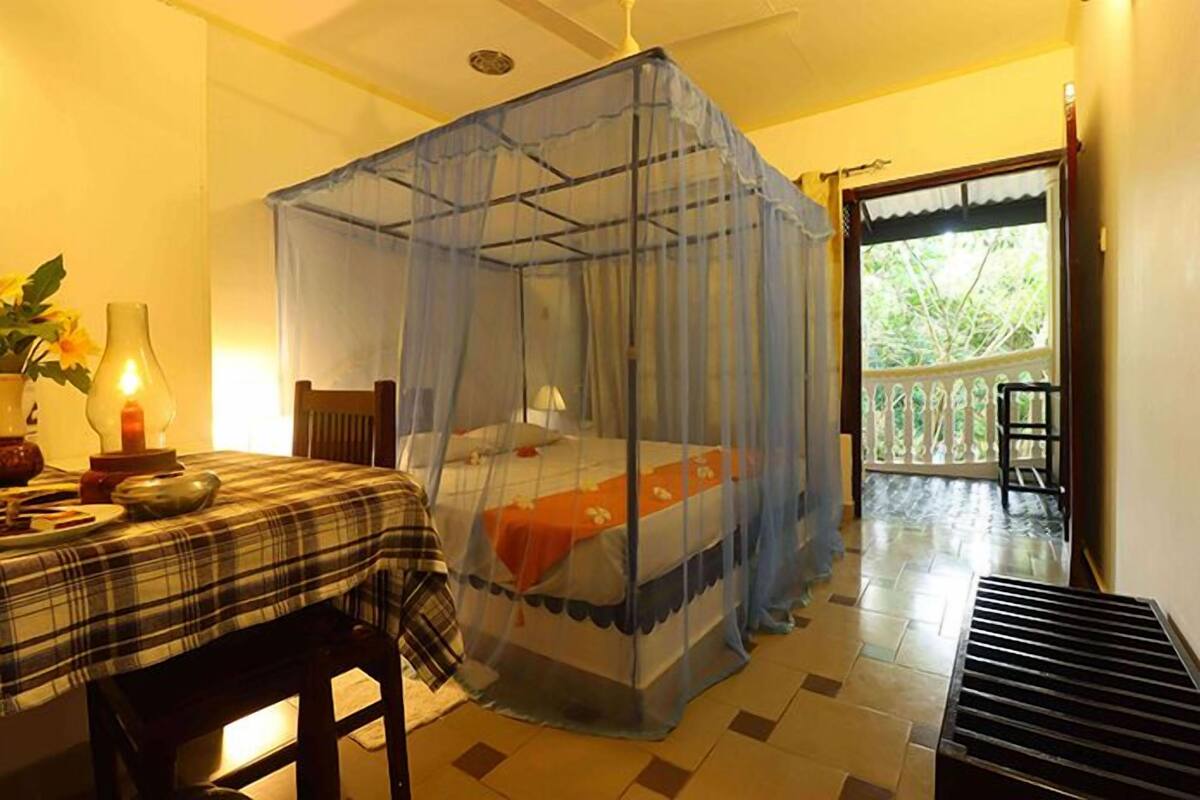 Een kamer met een bed onder een klamboe, een tafel met geruit tafelkleed en uitzicht op groen.