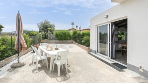 Une terrasse extérieure avec une table dressée pour le repas, entourée de verdure et d’un bâtiment blanc doté d’une porte-fenêtre coulissante.