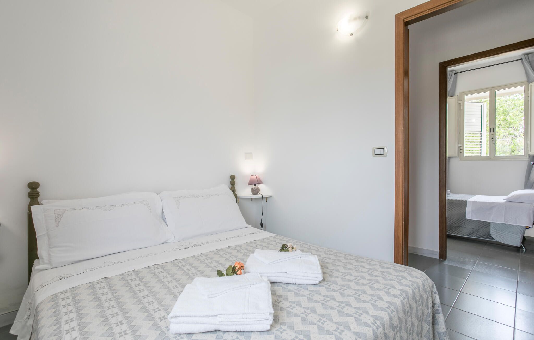 Quarto com cama, mesa de cabeceira e abajur, e banheiro visível por uma porta aberta.