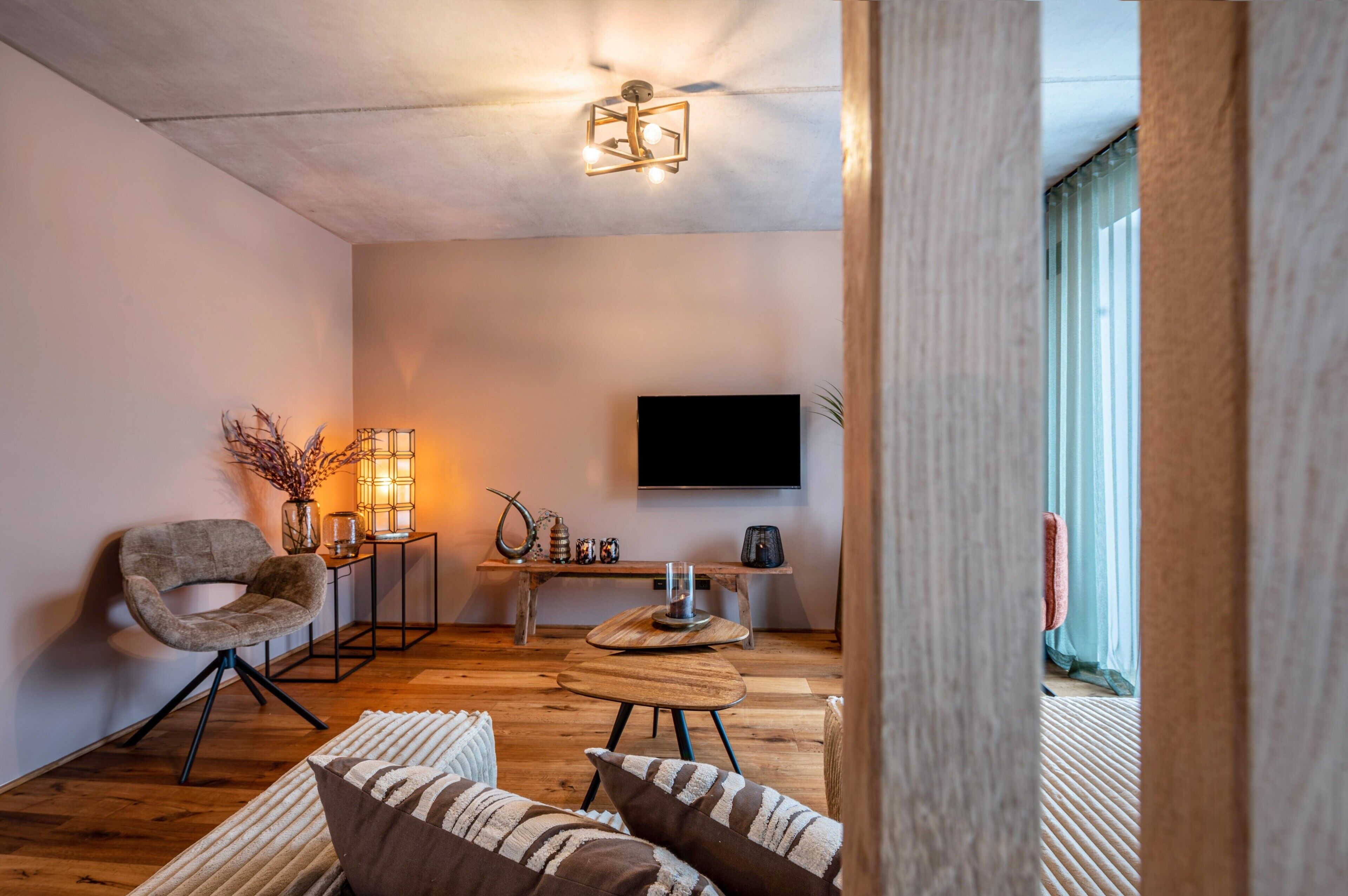 Ein modernes Wohnzimmer mit Holzboden, einem runden Couchtisch, einer Couch mit gestreiften Kissen, einem Sessel, einem an der Wand montierten Fernseher und einem kleinen Beistelltisch mit Dekoration.