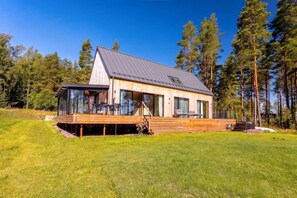 A modern wooden cabin with a large deck, surrounded by a forest.
