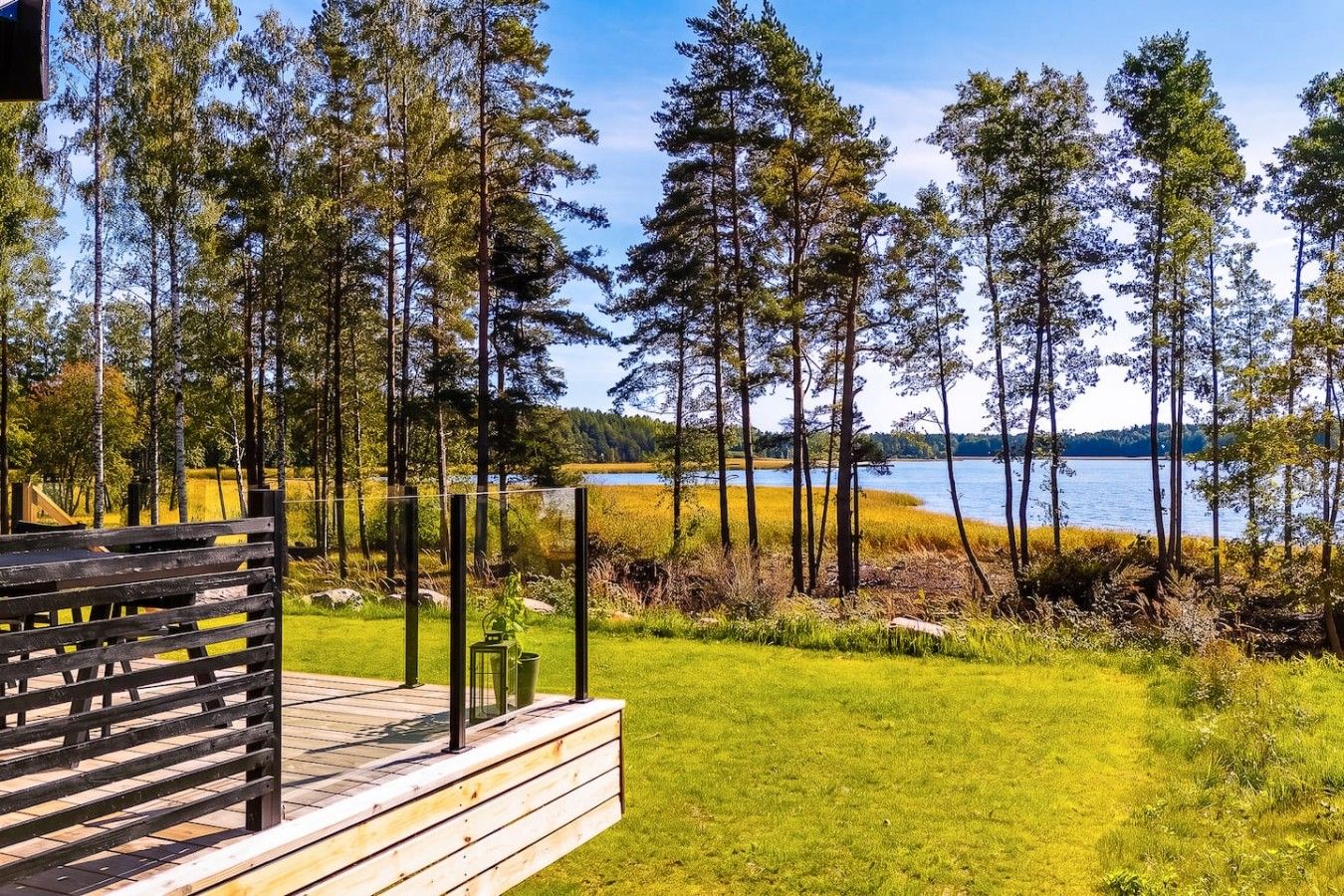 A wooden deck with a bench, overlooking a lake with tall trees and a grassy area.