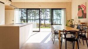 Une cuisine moderne dotée d’une grande fenêtre donnant sur un lac, un coin repas avec une table et des chaises en bois, et un aménagement intérieur minimaliste.
