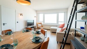 A modern living room with a wooden dining table set for a meal, a sofa, a TV, and a bookshelf. - Escape with sea view by Interhome (Blankenberge)