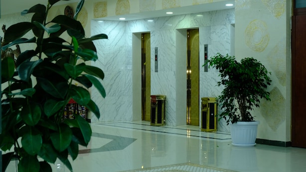 A lobby with marble flooring, a potted plant, and two elevators with gold doors. - Grand Bukhara (Bukhara)