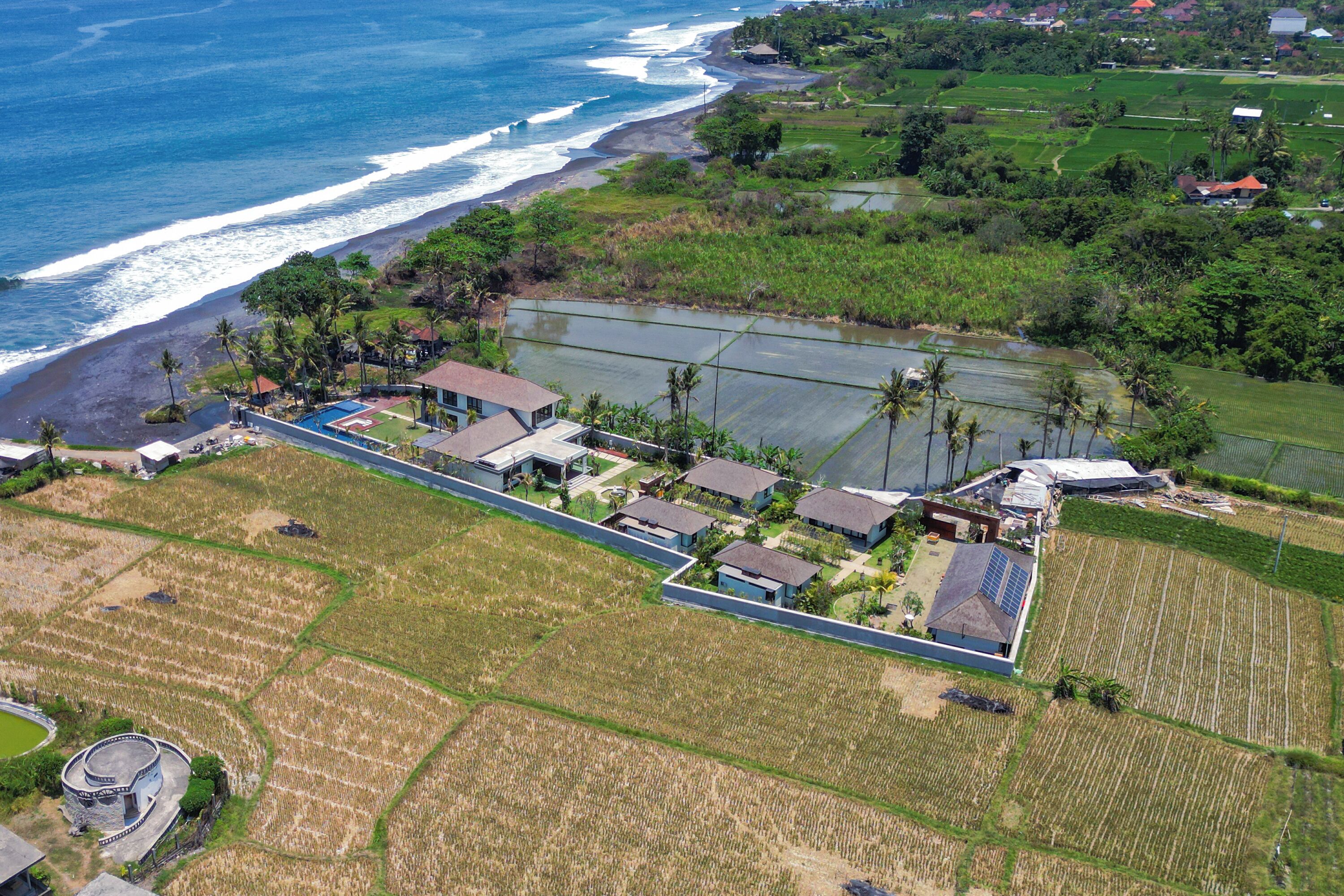 An aerial view of a coastal property with a long, narrow building complex adjacent to a beach, surrounded by agricultural fields and residential areas.