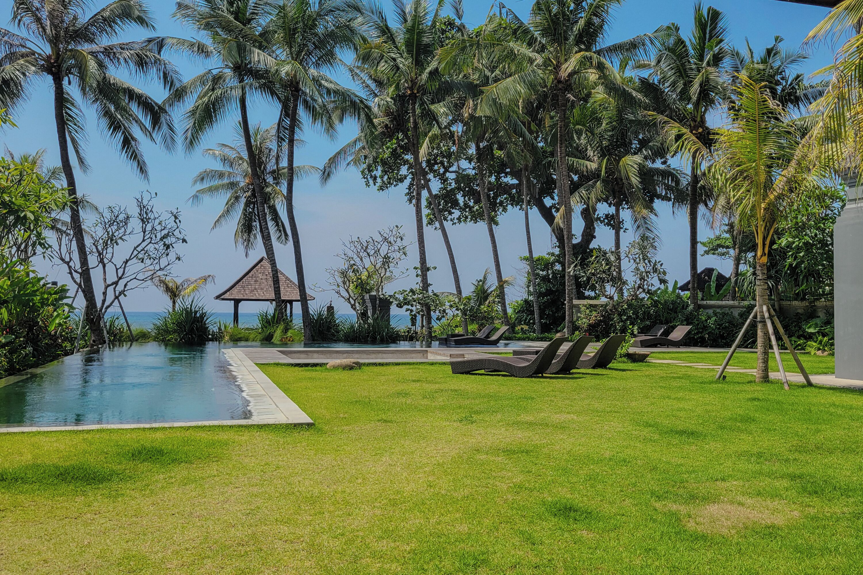 Zona junto a la piscina con sillones de descanso, palmeras y vista al océano.