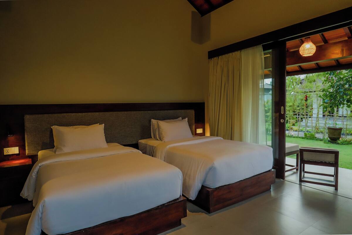 A hotel room with two beds, a wooden headboard, a small table, and a view of a garden through a glass door.