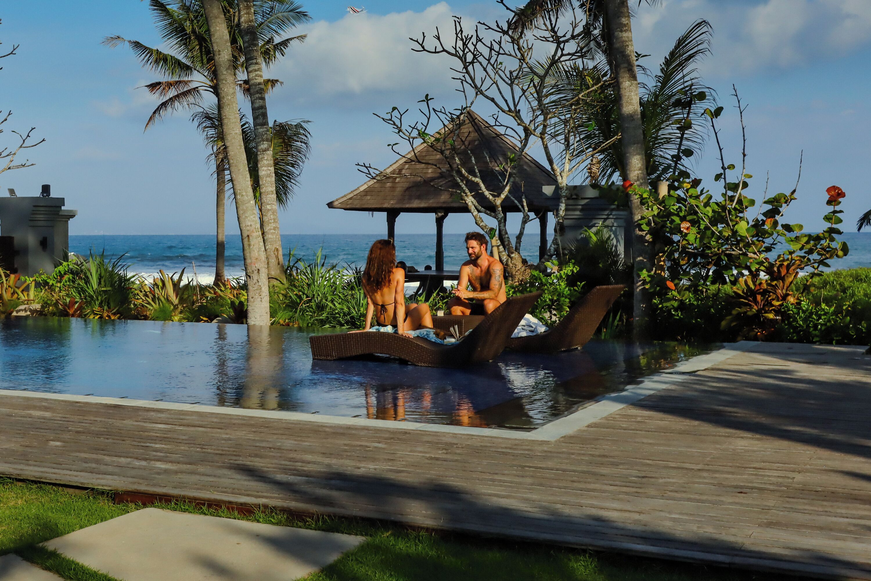 A couple relaxing by a pool with a view of the ocean, surrounded by tropical plants and a gazebo.