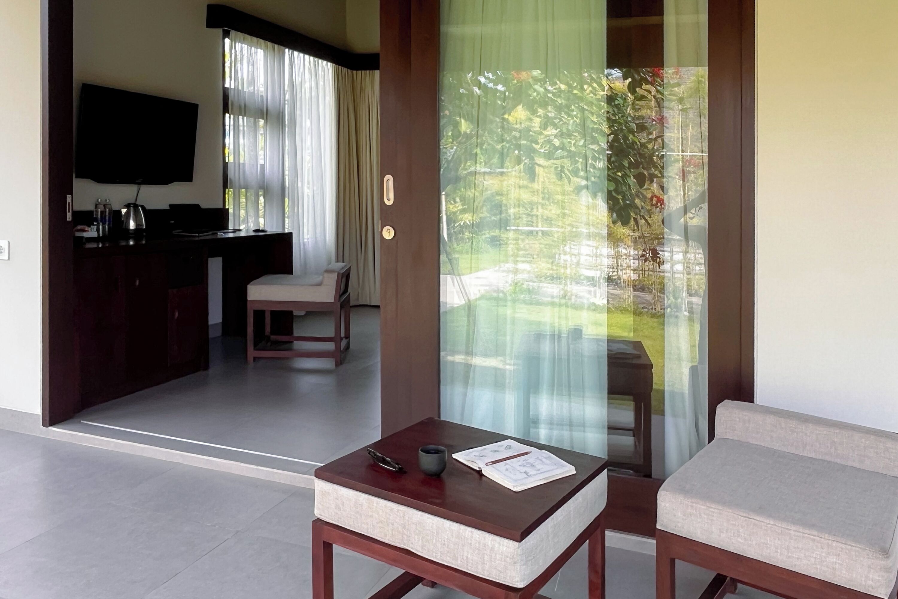 A modern room with a wooden table, a cushioned chair, a TV mounted on the wall, and a view of a garden through a glass door.