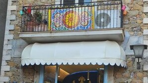 Un edificio de dos pisos con un balcón, una entrada a tienda con toldo azul y estatuas decorativas a cada lado.
