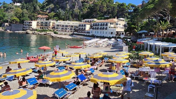 Una playa con varias tumbonas y sombrillas, un litoral rocoso y edificios al fondo.