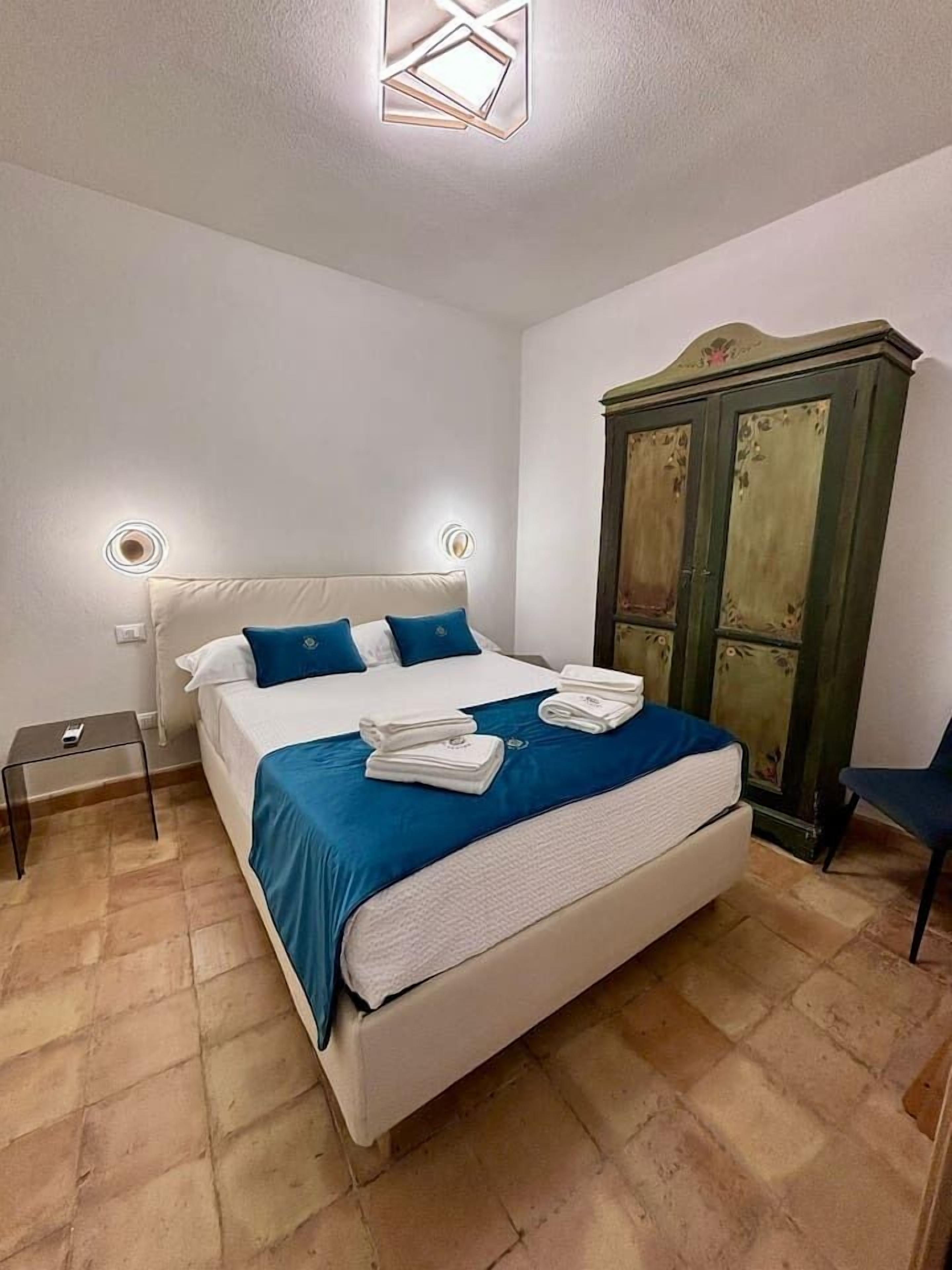 A neatly made bed with a blue bedspread, a wooden wardrobe, and a small bedside table with a lamp.