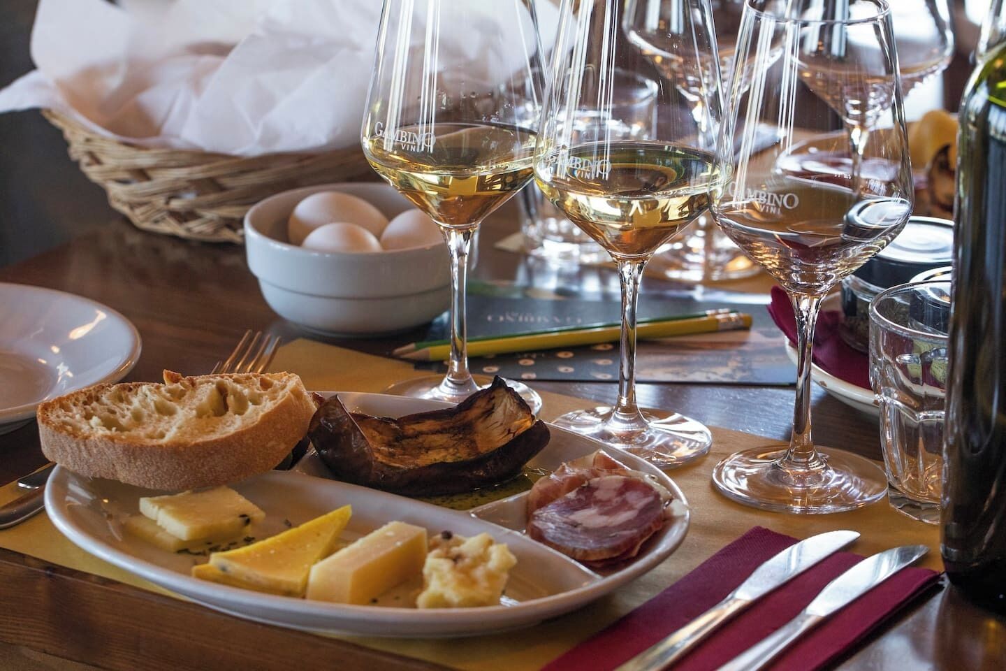 Une table à manger avec une assiette de nourriture, des verres à vin et un panier d’œufs.