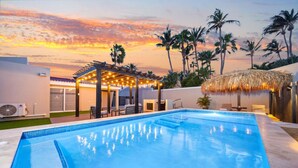 Uma piscina com pergolado e guarda-sol de palha, rodeada por palmeiras e vista para o pôr do sol.