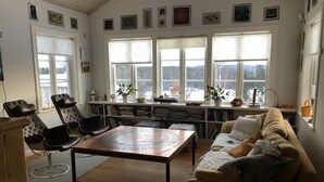 Un salón acogedor con una mesa de centro de madera, un sofá color beige y un sillón de cuero negro. Se observan ventanas grandes con cortinas, una estantería llena de libros y una pequeña mesa con adornos.