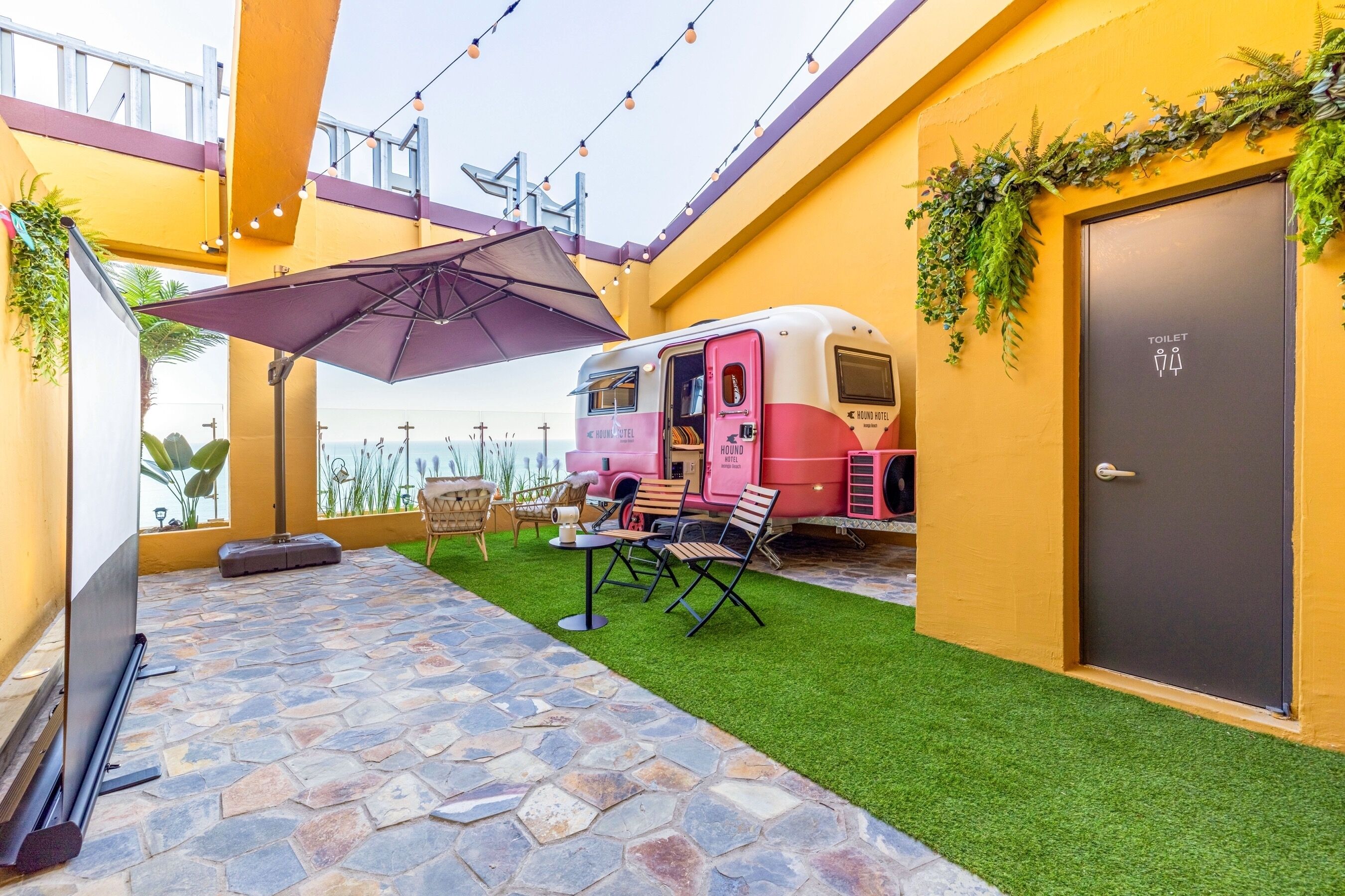 Un espace patio doté d’un sol en pierre, d’une pelouse artificielle et d’une pièce de style caravane rose, avec un panneau indiquant les toilettes.