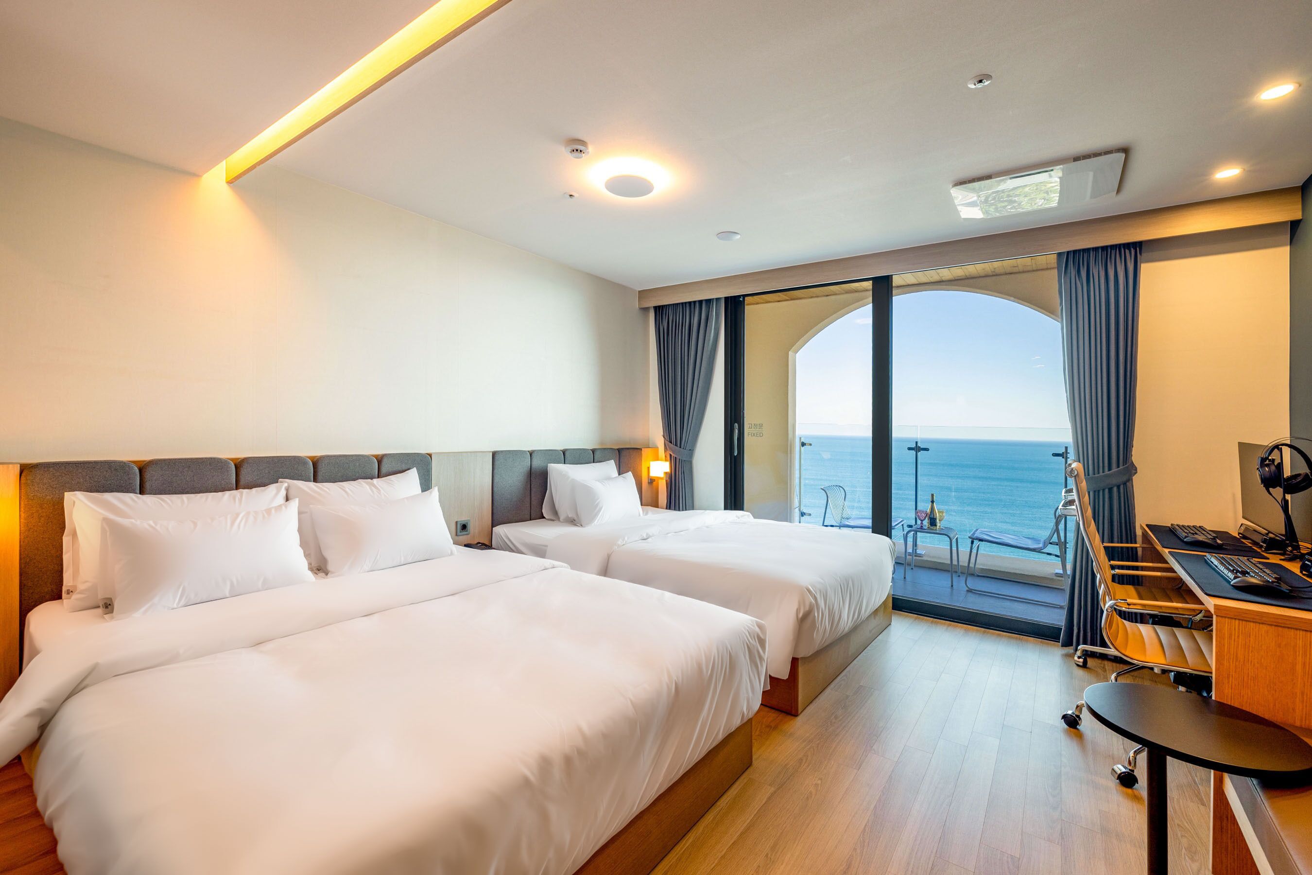 A hotel room with two beds, a desk, and a view of the sea.
