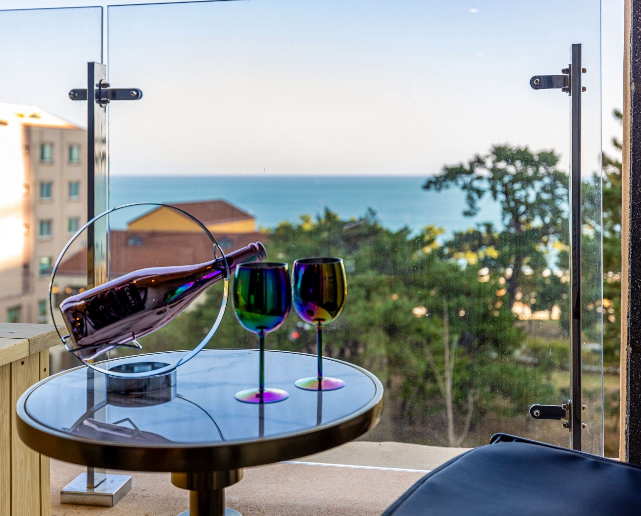 A table with a pair of sunglasses and two wine glasses, overlooking a view of the sea and buildings.