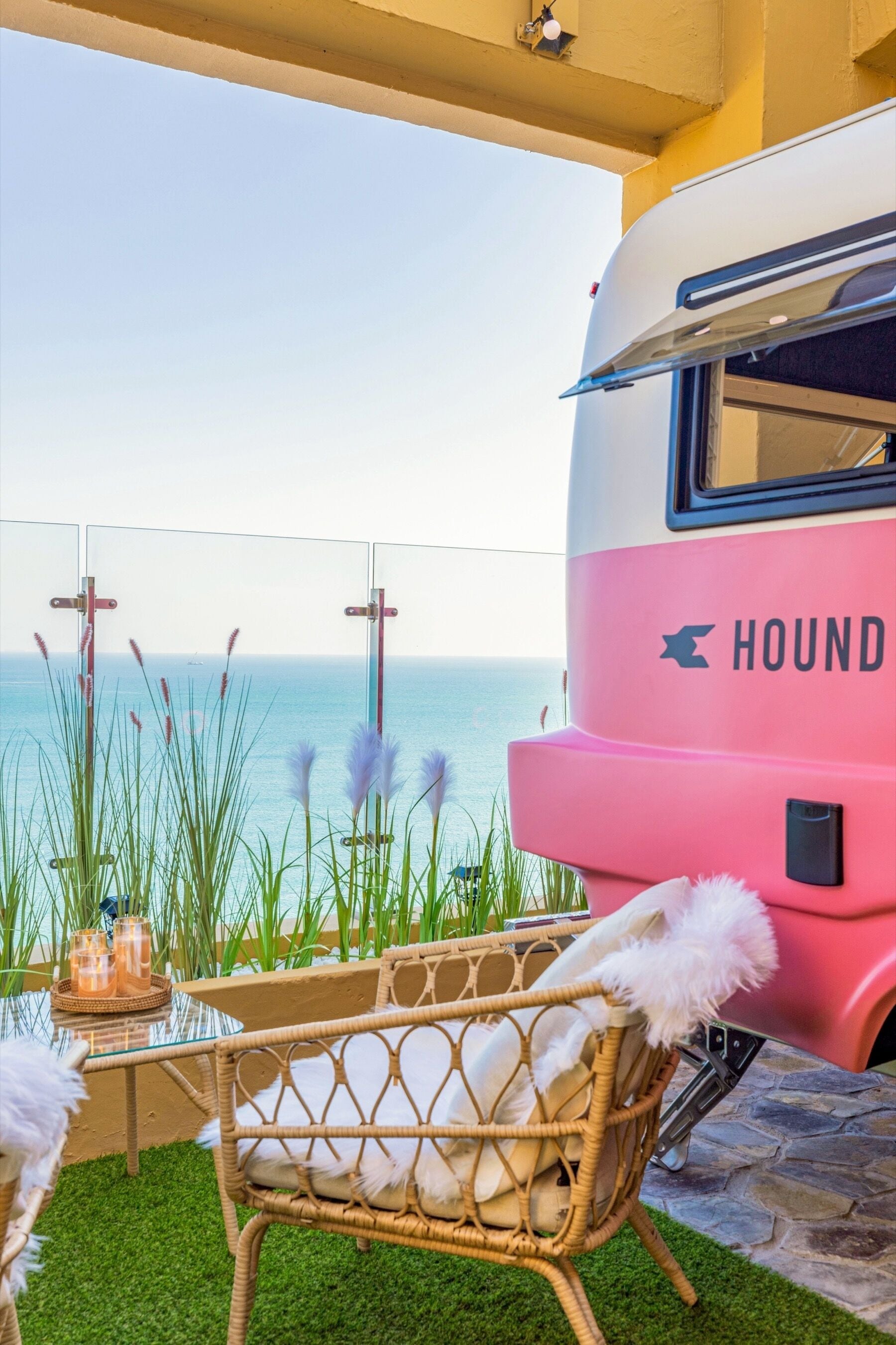 Una caravana rosa con la palabra "HOUND" en el lateral estacionada en una zona costera, con una silla de mimbre y una mesa de vidrio con bebidas.