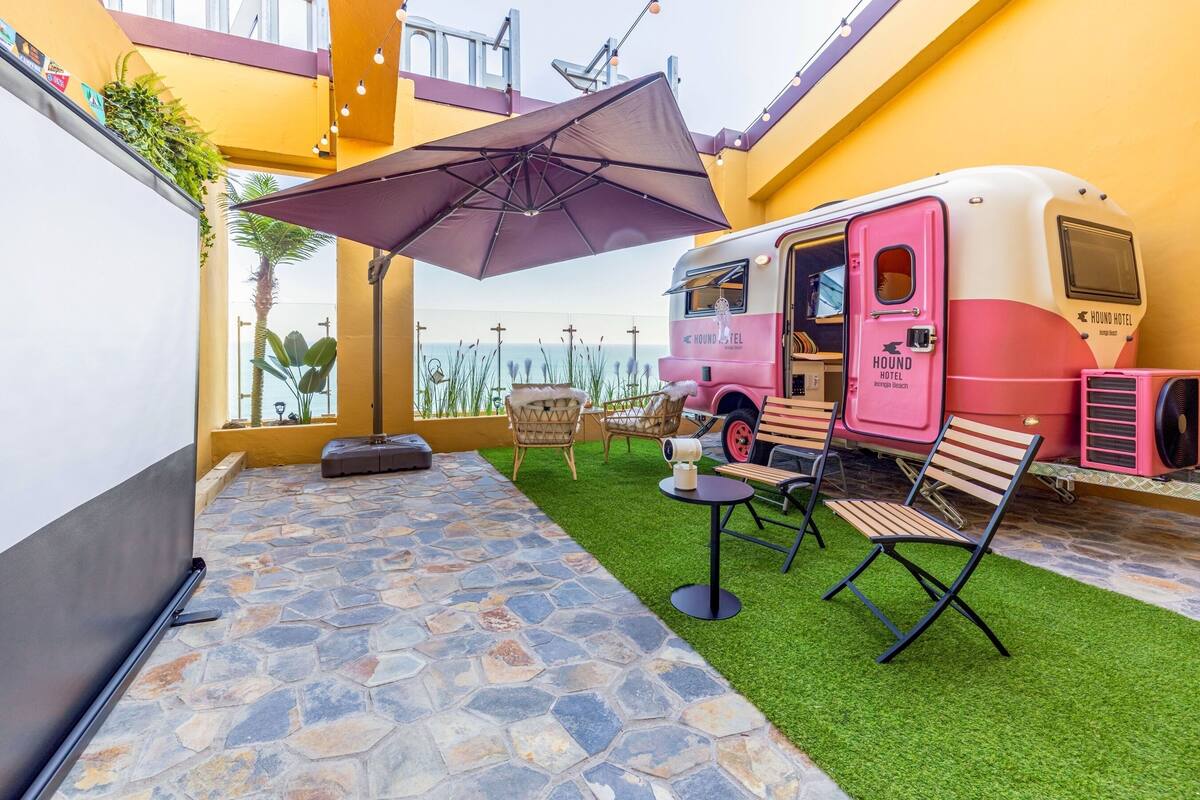 A dining area with a pink RV-style food truck, outdoor seating, and a stone-tiled floor.