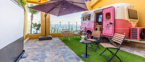 A dining area with a pink RV-style food truck, outdoor seating, and a stone-tiled floor.