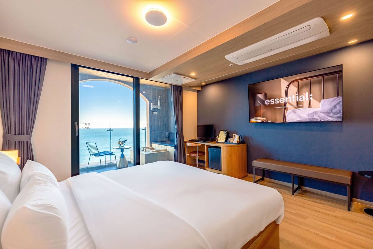 A hotel room with a large bed, a desk with a chair, a TV, and a view of the sea.