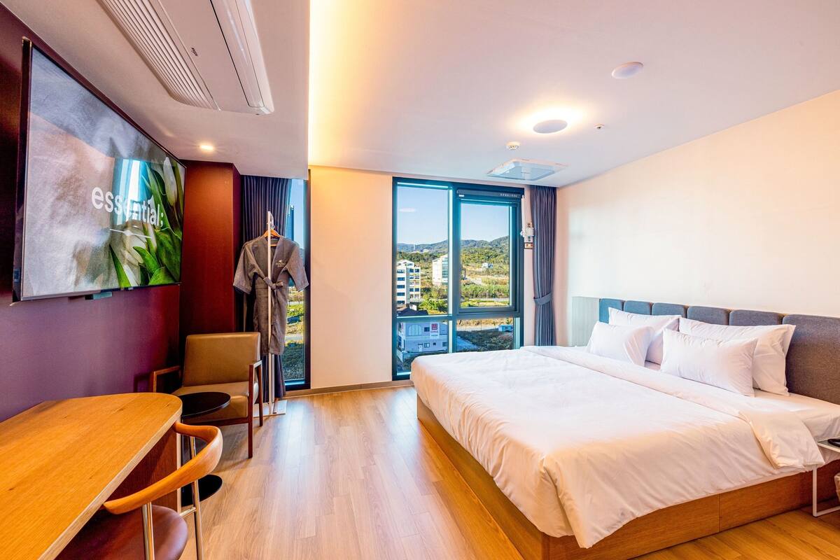 A modern hotel room with a large bed, a desk, and a view of the city through a glass door.