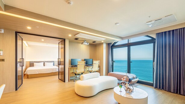 A modern hotel room with a large bed, a seating area with a coffee table, a desk with a chair, and a large window offering a view of the sea. - Hound Hotel Ulsan (Ulsan)
