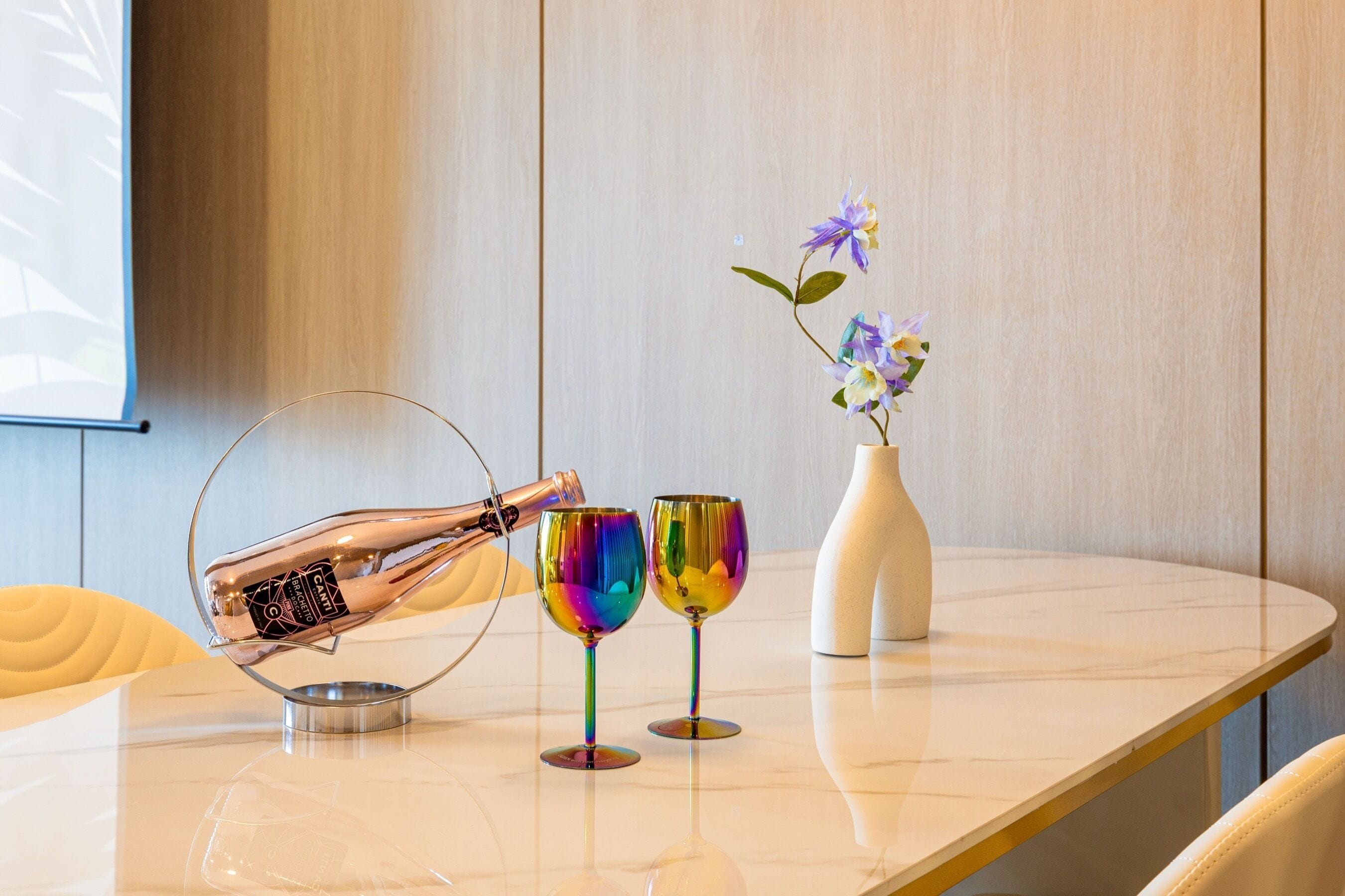 A table with two wine glasses, a vase with flowers, and a champagne bottle on a marble surface.