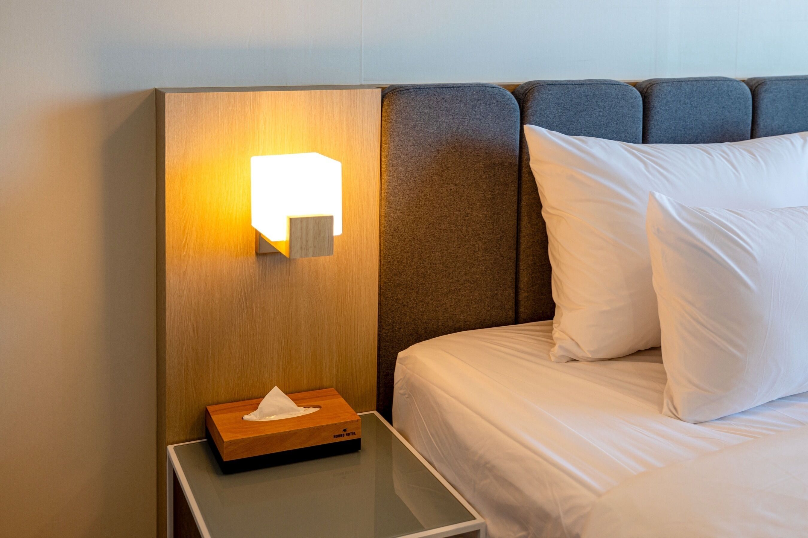 A hotel room with a neatly made bed, a bedside table with a tissue box, and a wall-mounted light fixture.