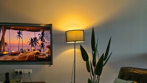 A living room with a television displaying a beach scene, a floor lamp, a sofa with a patterned cushion, and a potted plant.