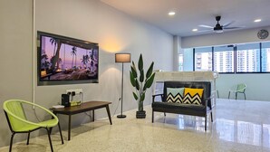A modern living room with a large flat-screen TV, a sofa with patterned cushions, a green chair, a floor lamp, and a potted plant.