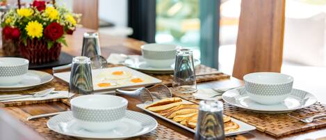 A breakfast setting with a table set for two, including plates, bowls, cups, and a basket of flowers.