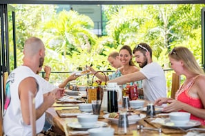 Un grupo de personas cenando en una mesa al aire libre, con diversos platos y bebidas, rodeado de abundante vegetación.