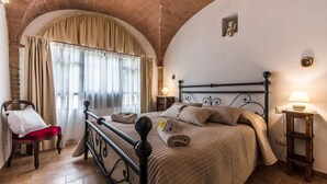 Ein Schlafzimmer mit Ziegelbogen, einem Bett mit Kopfteil, zwei Nachttischen mit Lampen, einem Sessel und einem Fenster mit Vorhängen.