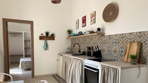 Una cocina compacta con estufa, horno y lavamanos, una pequeña zona de comedor con una silla, y un pasillo que conduce a un dormitorio.