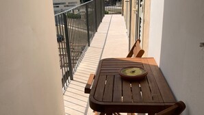 Un balcón estrecho con una mesa y una silla de madera, una planta en maceta y vistas a edificios al fondo.