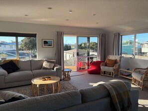 A modern living room with a grey sofa, a red chair, and a wicker chair. There is a wooden coffee table and a view through large windows. - Hosts on the Coast - Catch of the Day (Cooks Beach)