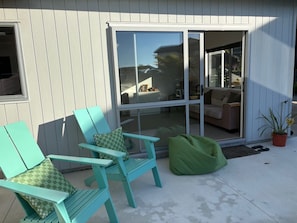 Une terrasse avec deux chaises turquoise et un pouf vert, une porte-fenêtre coulissante donnant sur un espace de vie intérieur, et une plante en pot posée sur le sol.
