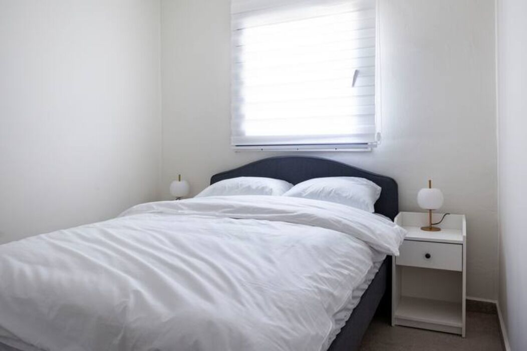 A bedroom with a bed, white bedding, a nightstand with a lamp, and a window with blinds.