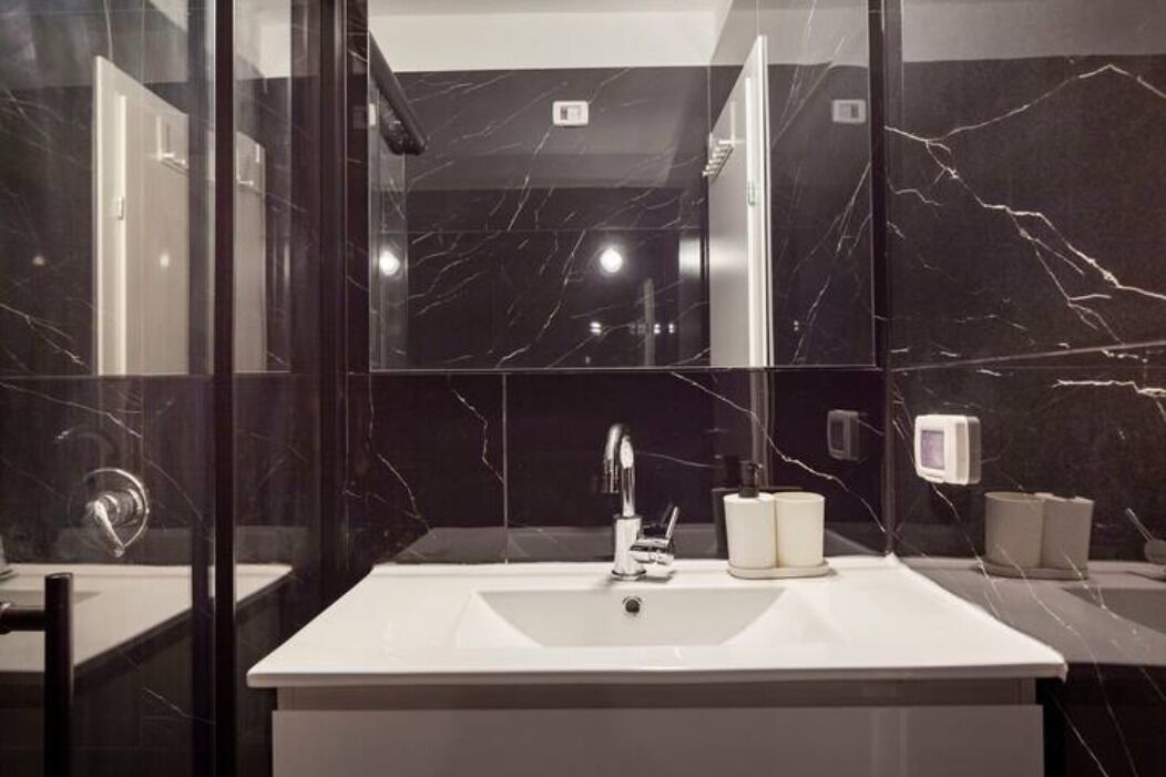Un baño moderno con un lavamanos blanco, paredes de mármol oscuro y cabina de ducha de vidrio.
