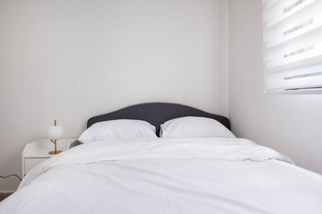 A neatly made bed with white linens and a dark headboard, a small bedside table with a lamp, and a window with blinds.