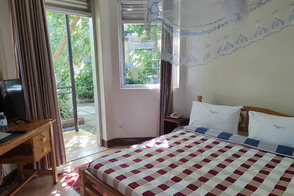A hotel room with a bed, a desk with a TV, a window with curtains, and a view of greenery outside.