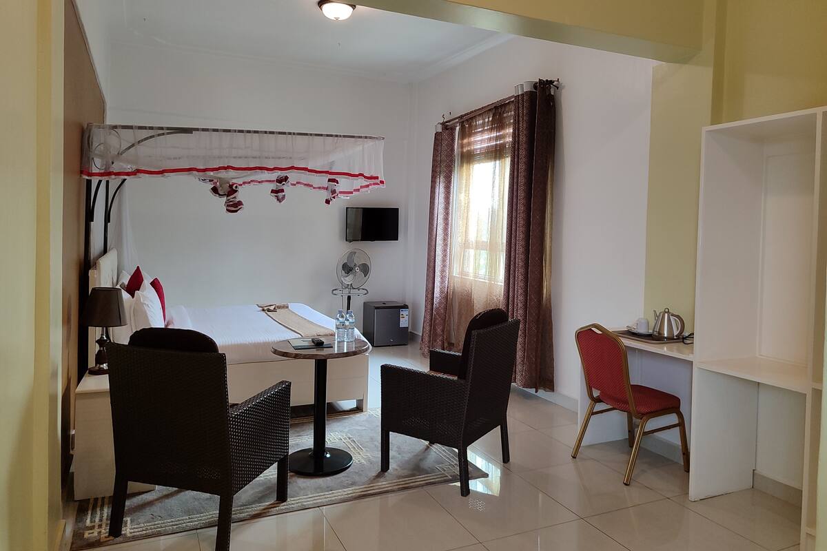 A hotel room with a bed, a small table with chairs, a desk with a chair, a TV, and a window with curtains.