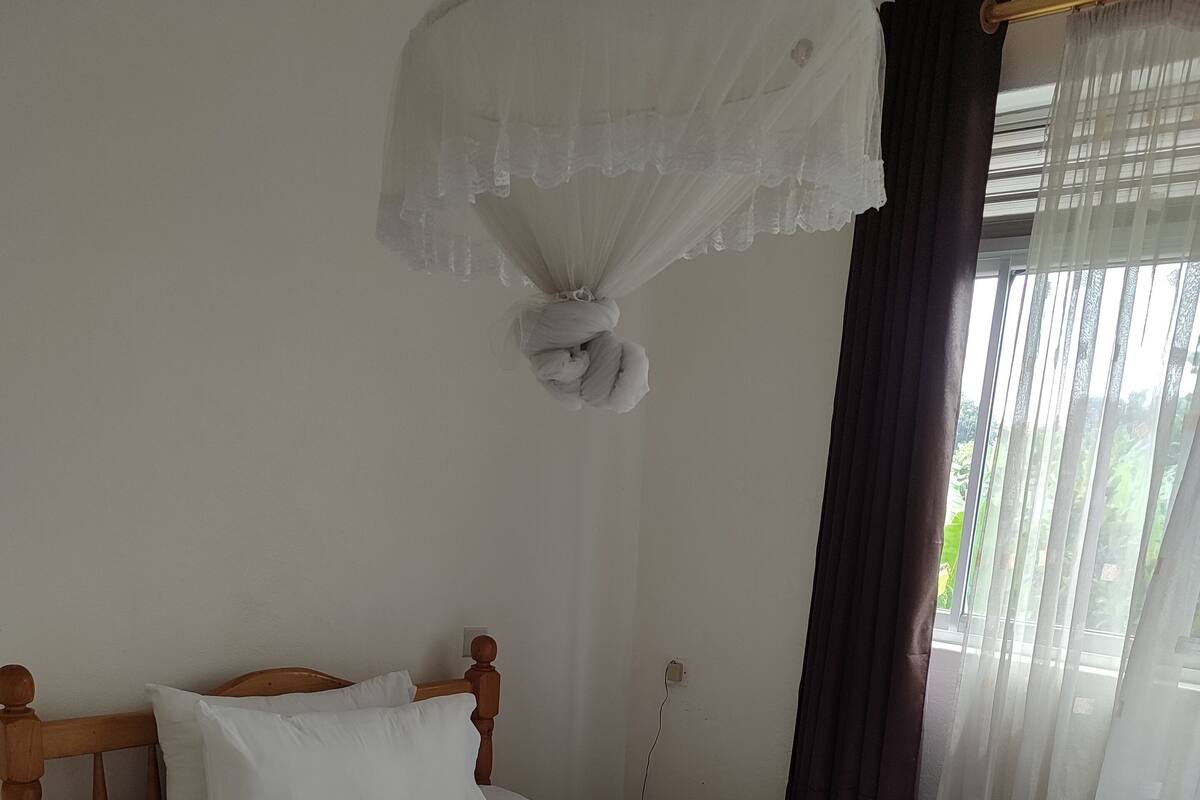 A neatly made bed with a wooden frame and white bedding, a power outlet on the wall, and a window with sheer curtains.